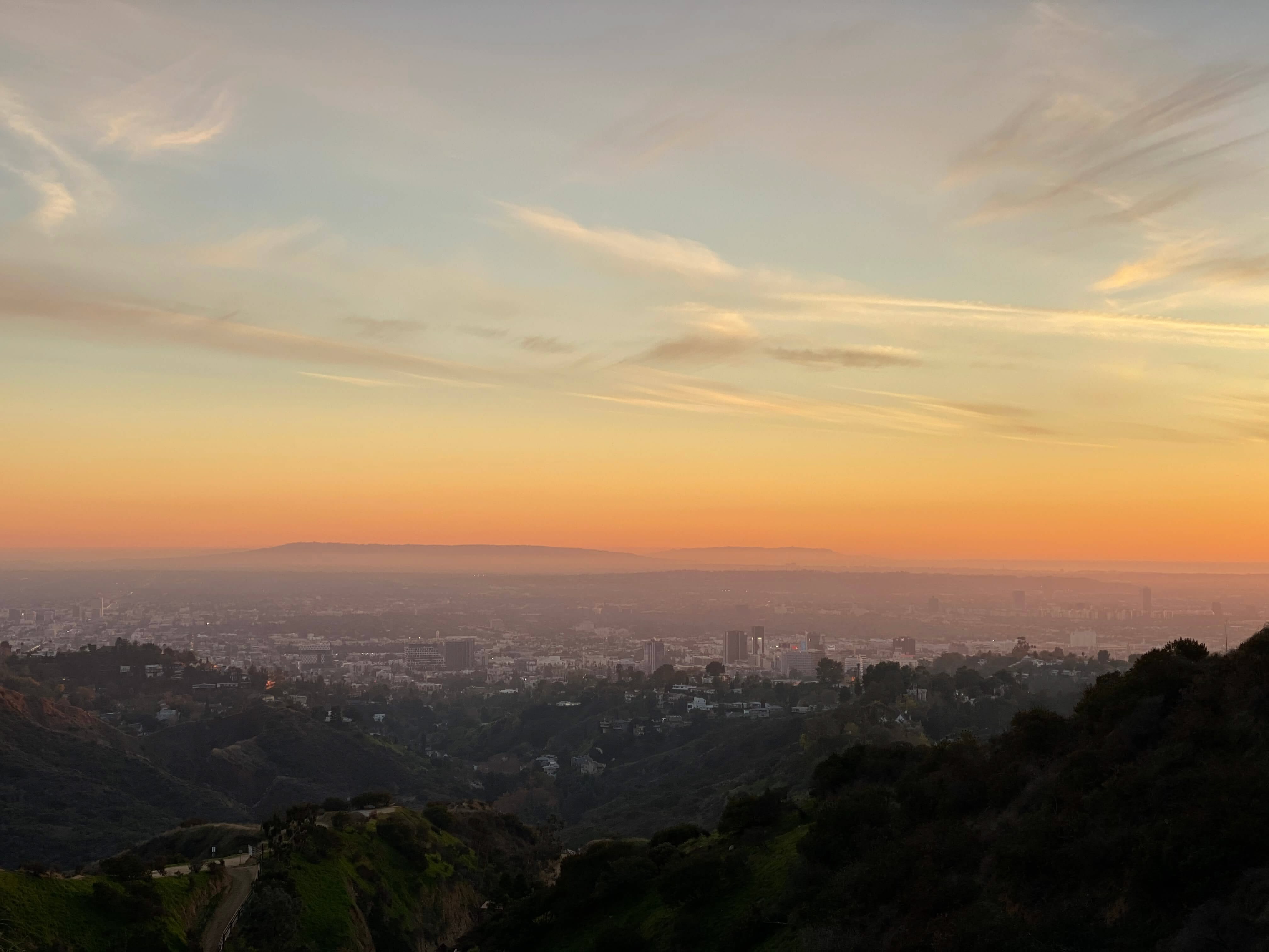 Sunset view over Los Angeles from elevated hillside with wispy clouds painted in orange and blue, city sprawling below with dark vegetation in foreground